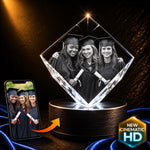 A diamond-shaped acrylic photo lamp on a cylindrical base, displaying a black-and-white graduation photo of three women. A smartphone on the left shows the source photo, with an orange arrow indicating the projected image.